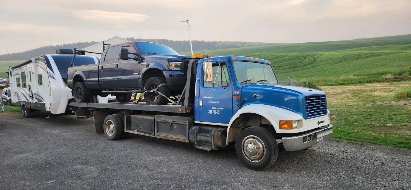 Snoway Customs blue flatbed tow truck hauling a pickup and travel trailer across the Palouse